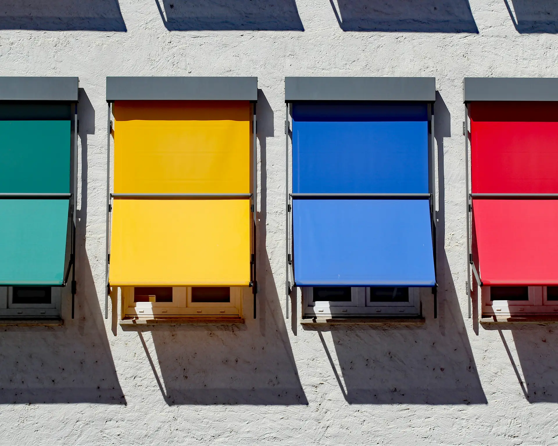 Vier Fenster mit bunten Rollläden in Grün, Gelb, Blau und Rot an einer weißen Fassade